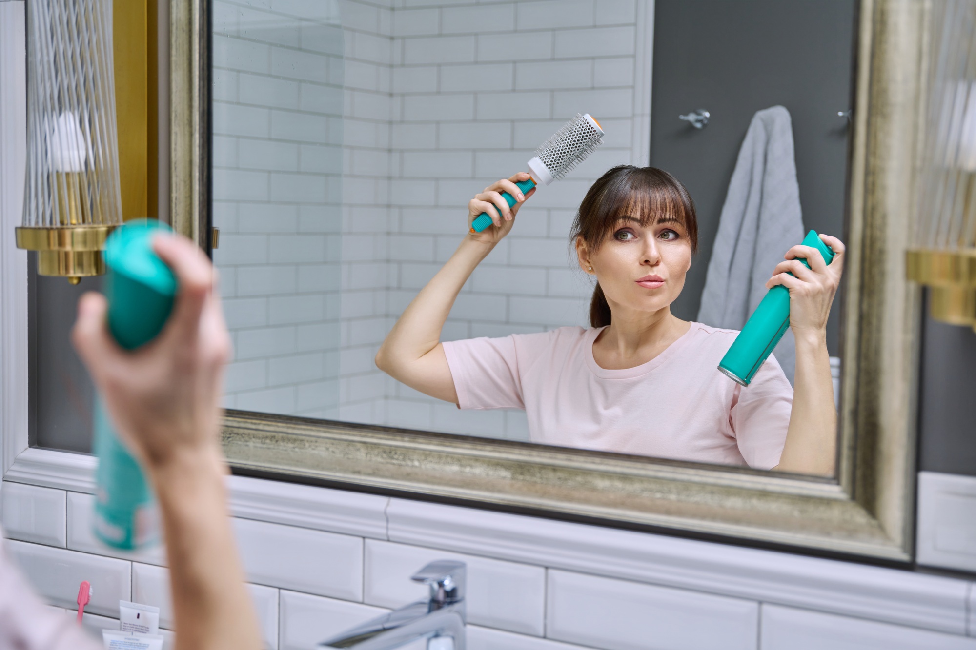 Middle-aged woman with hairspray comb looking in mirror, doing hair styling, in bathroom